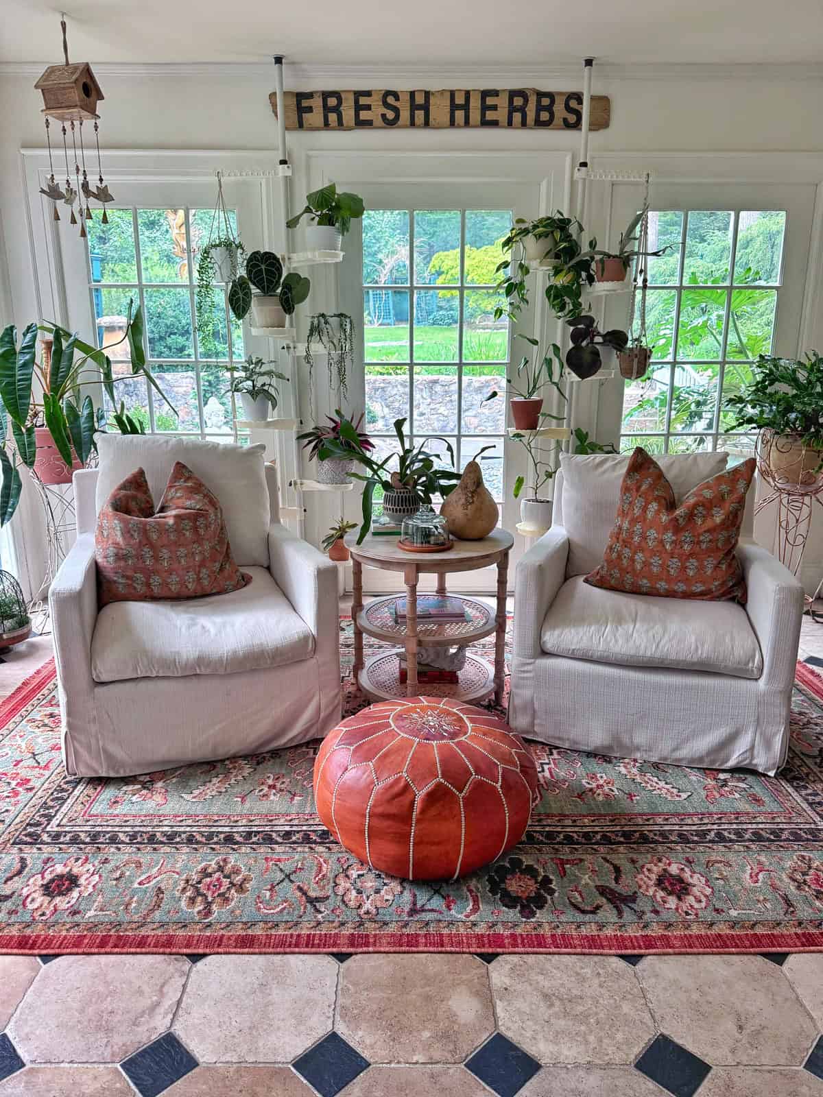 Two white armchairs with patterned pillows sit on a colorful rug, separated by a small table with plants. Greenery and hanging plants decorate the bright sunroom. A sign above the windows reads “FRESH HERBS.”.