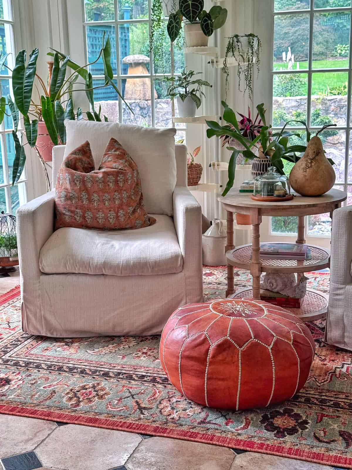 A cozy sunroom with a beige armchair, decorative pillows, a round orange pouf, a patterned rug, side table with plants and decor, and large windows showing a green outdoor view.