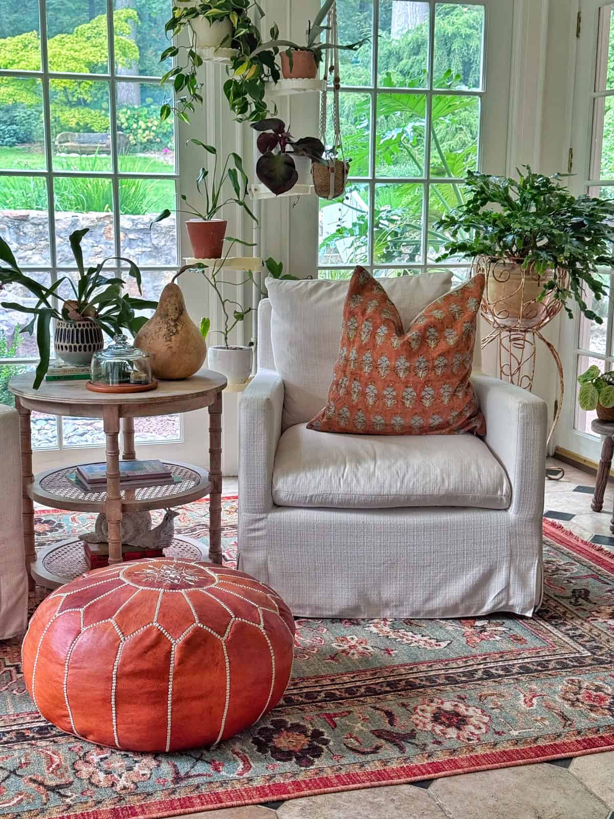 A cozy white armchair with an orange patterned pillow sits by tall windows, surrounded by plants. An orange pouf and a small side table with pottery rest on a colorful rug. Lush greenery is visible outside.