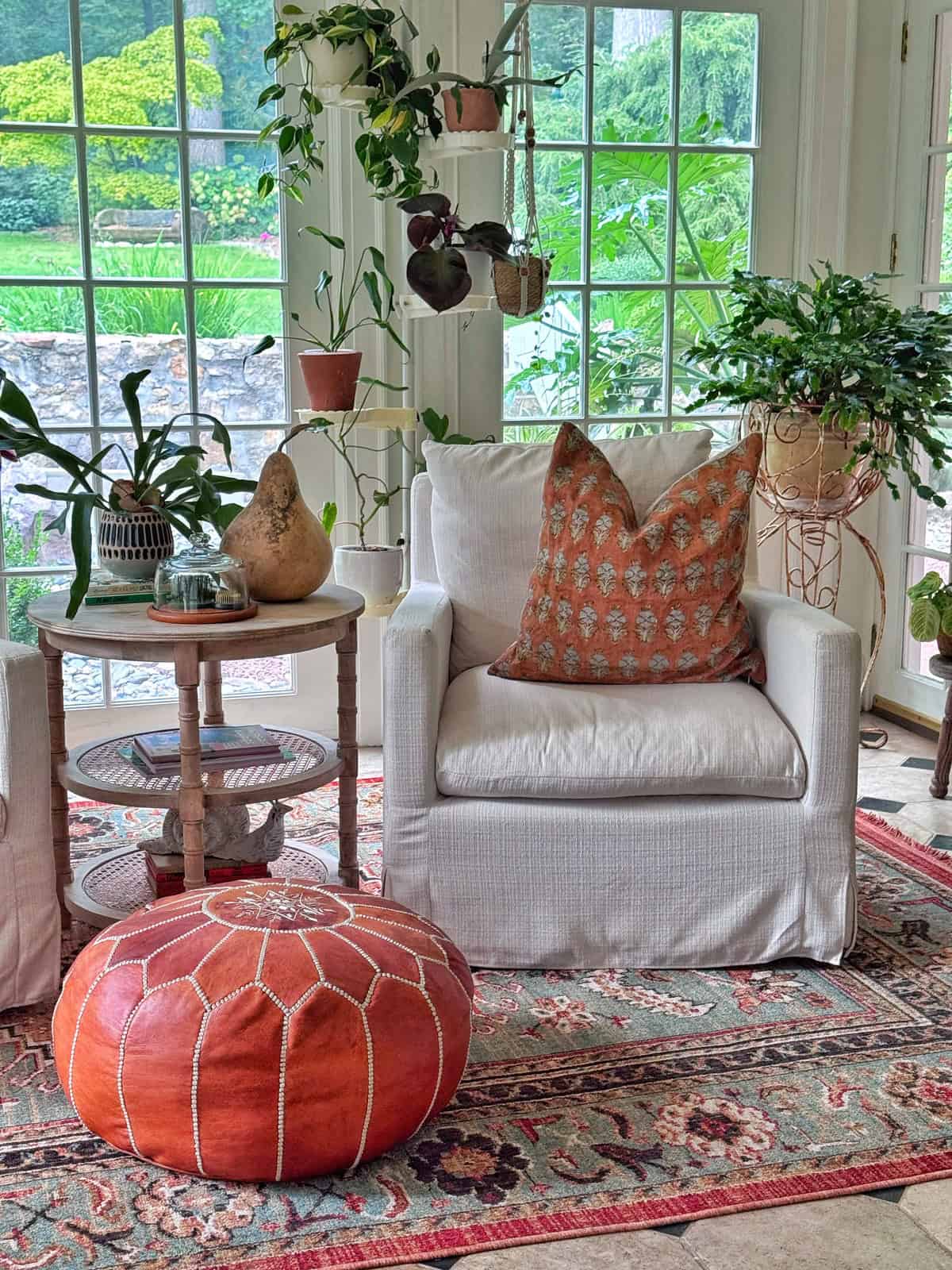 A cozy sunroom with a white armchair, orange pillow, round leather pouf, side table with plants and decor, patterned rug, and potted plants by large windows overlooking a green outdoor scene.