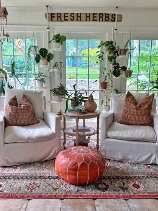 Cozy sunroom with two white armchairs, patterned pillows, a round leather pouf, hanging and potted plants, a side table with decor, and a “FRESH HERBS” sign above large windows overlooking a lush garden.