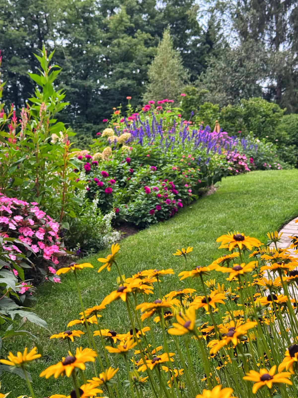 A lush garden with vibrant yellow, pink, purple, and red flowers borders a neatly trimmed green lawn, with tall trees in the background on a cloudy day.