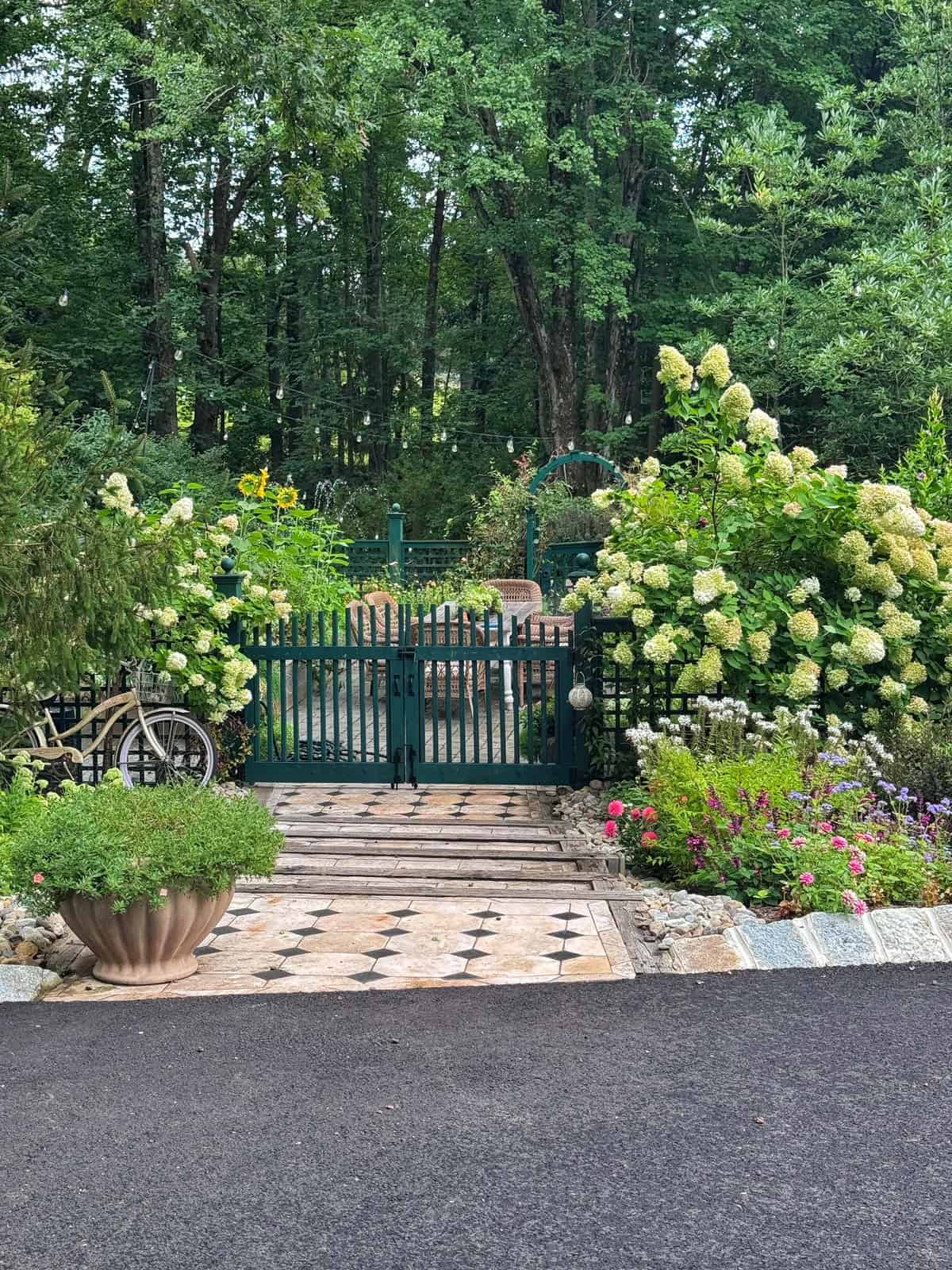 A black metal gate opens to a lush garden with blooming hydrangeas, stone paths, a bicycle leaning on the fence, and a large planter. Dense woods form the backdrop.