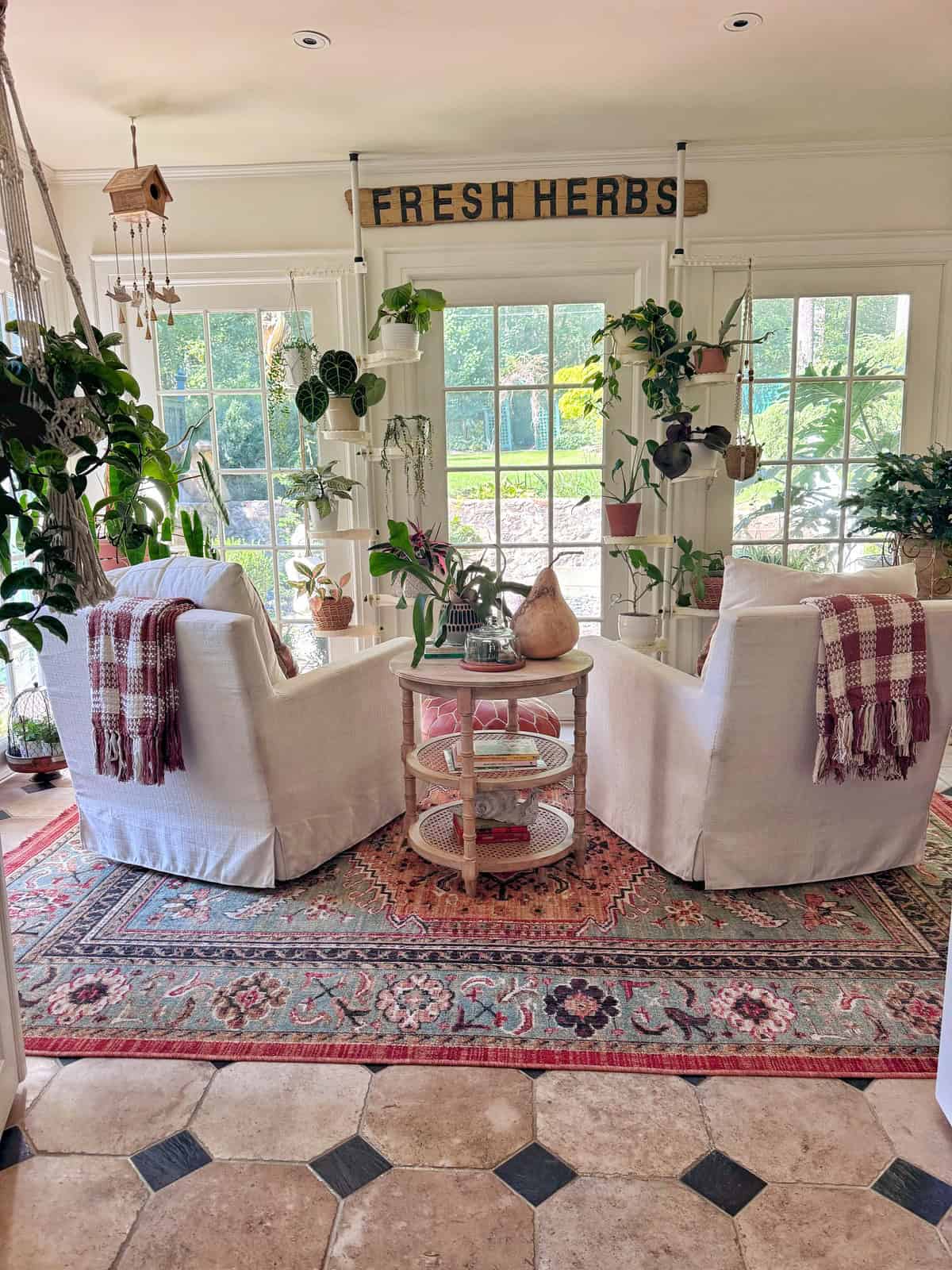 Two white armchairs with plaid blankets face each other by a round table in a bright, plant-filled sunroom. A “FRESH HERBS” sign hangs above large windows overlooking a garden.