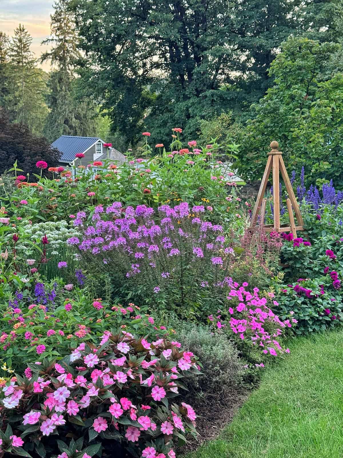 A lush garden with pink, purple, and red flowers, green shrubs, a wooden garden obelisk, and a small house in the background surrounded by tall trees.