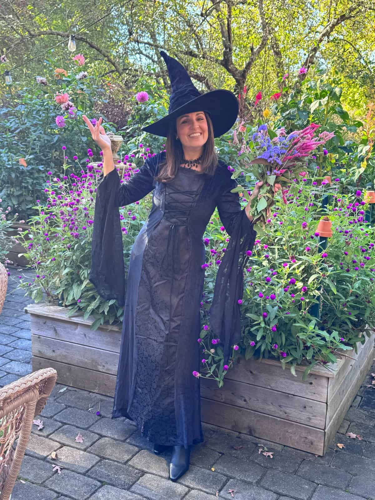A woman wearing a black witch costume and hat stands in a garden holding a bouquet of purple and pink flowers.