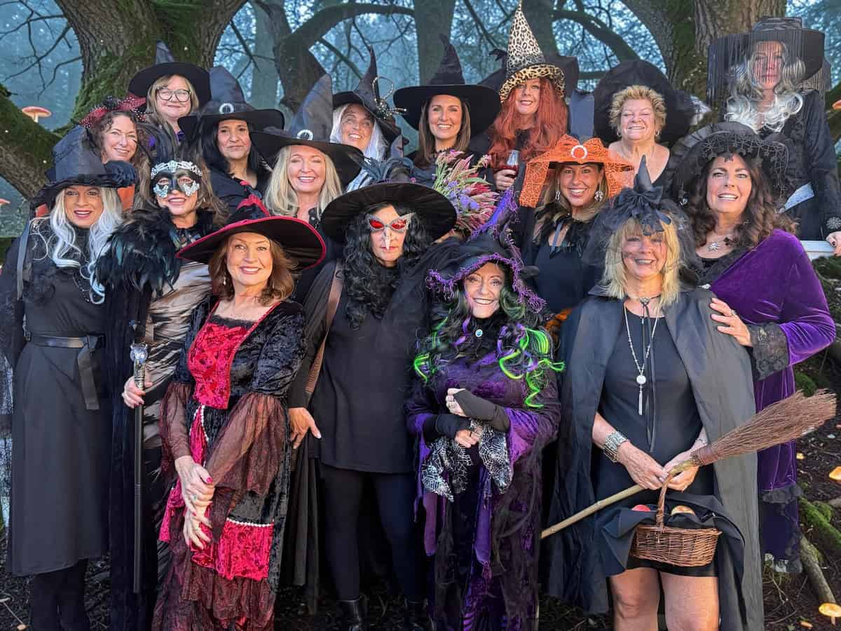 A group of women in various witch costumes and pointed hats posing for a photo outdoors in a wooded area.