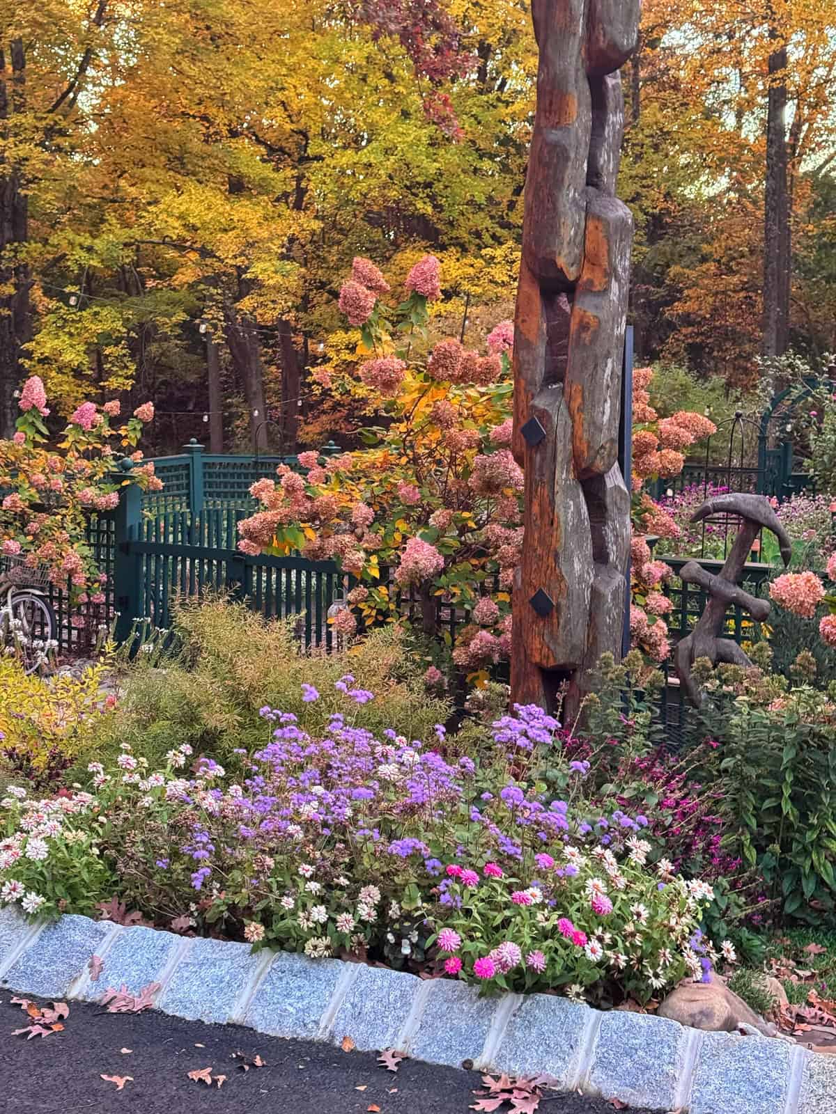 A vertical wooden sculpture of interlocking links stands in a garden filled with purple flowers, pink hydrangea bushes, and autumn trees.