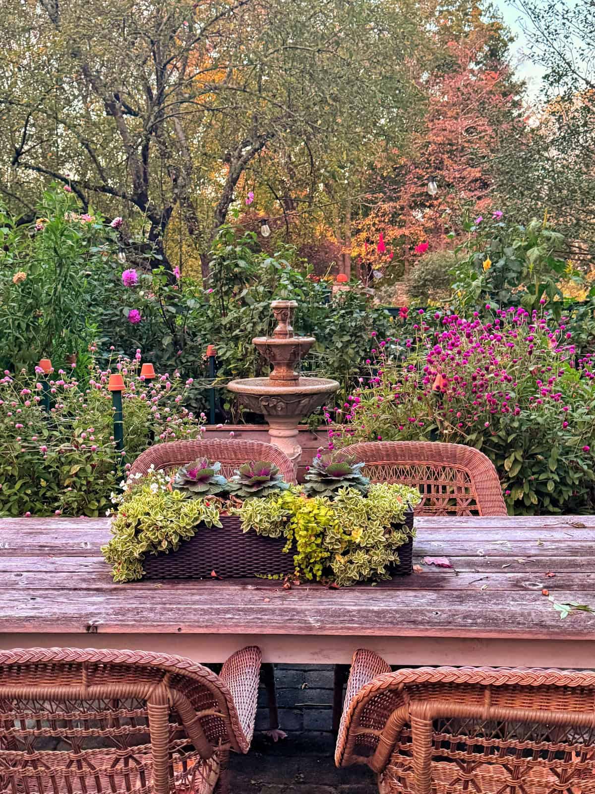 A weathered wooden table with wicker chairs sits in a garden in front of a stone fountain surrounded by flowers and trees.