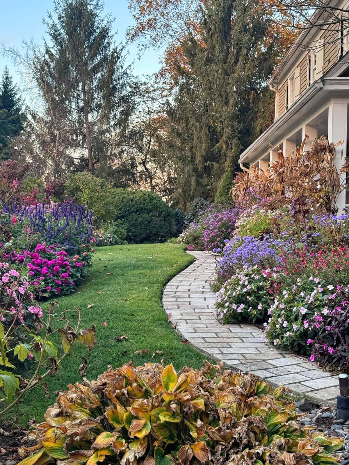 A winding stone path leads through a lush garden filled with colorful purple, pink, and white flowers beside a house porch.