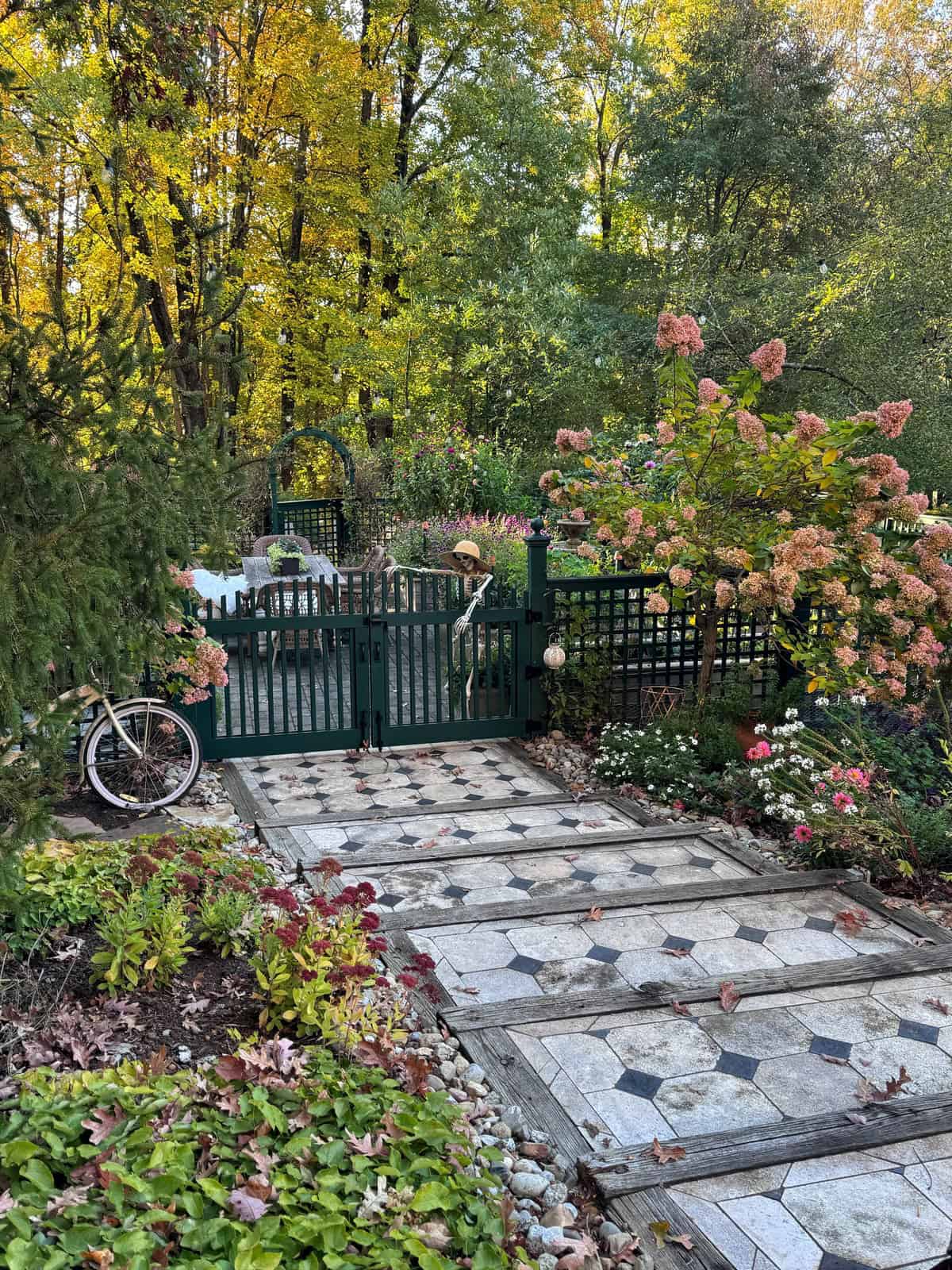 A patio area behind a dark green gate, featuring a tiled path, a vintage-style bicycle, and fall foliage in a garden setting.
