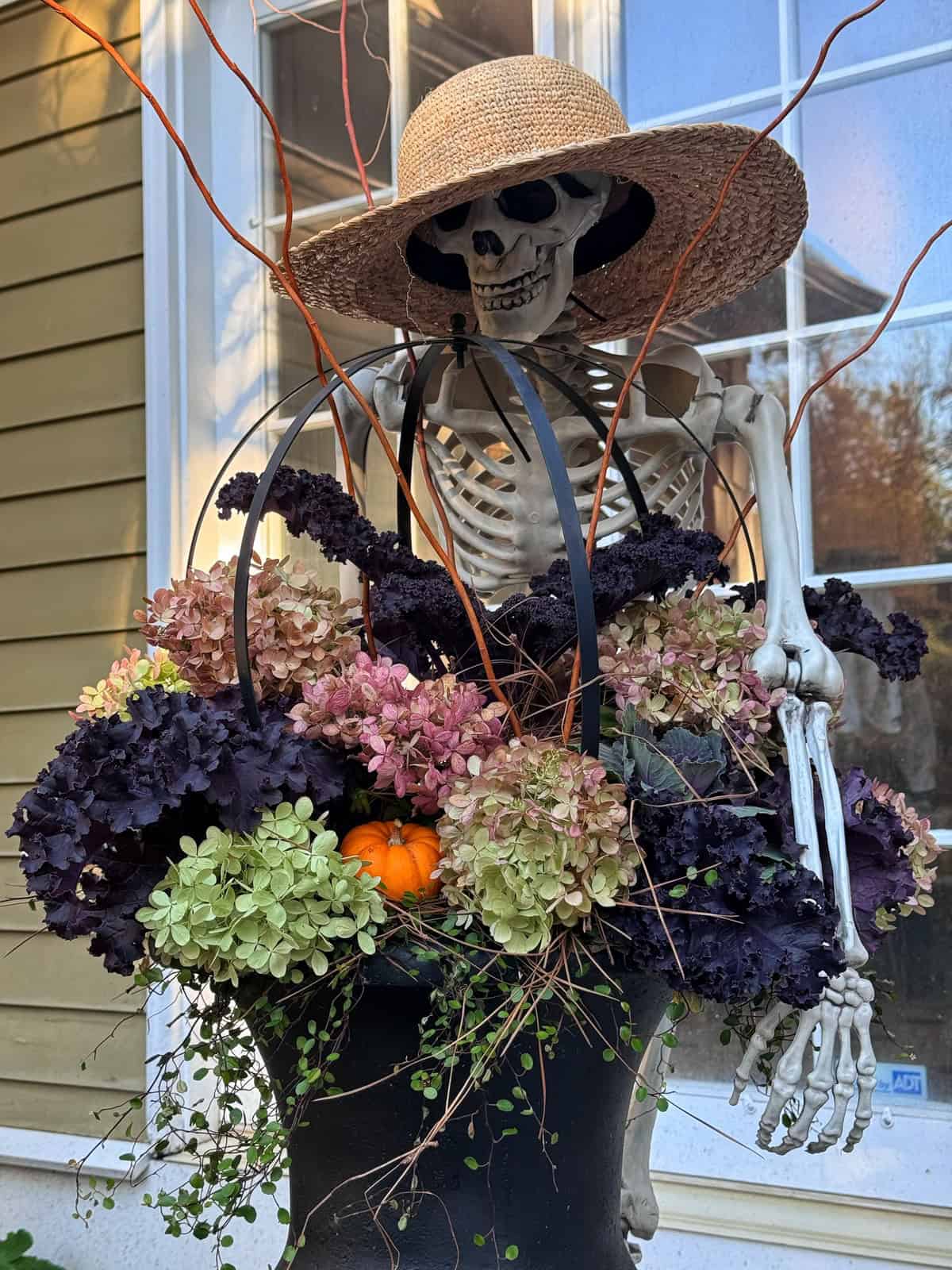 A skeleton wearing a straw hat sits behind a large planter filled with hydrangeas, purple kale, and a small pumpkin.