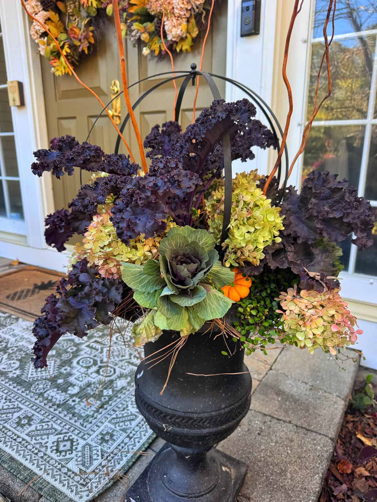 A black pedestal planter filled with purple kale, green ornamental cabbage, light green hydrangeas, thin orange branches, and a small decorative pumpkin.
