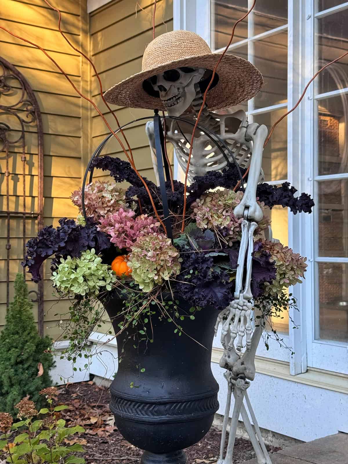 A skeleton wearing a straw hat stands next to a large black urn overflowing with hydrangeas, kale, and a small decorative pumpkin.