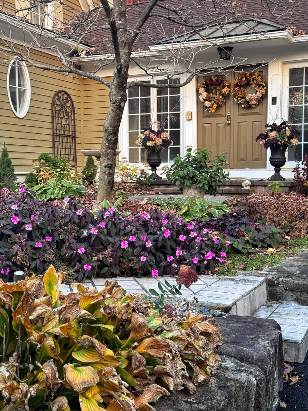 A front entryway with a brown door decorated with two fall wreaths, surrounded by autumn foliage, flowers, and a stone garden bed.